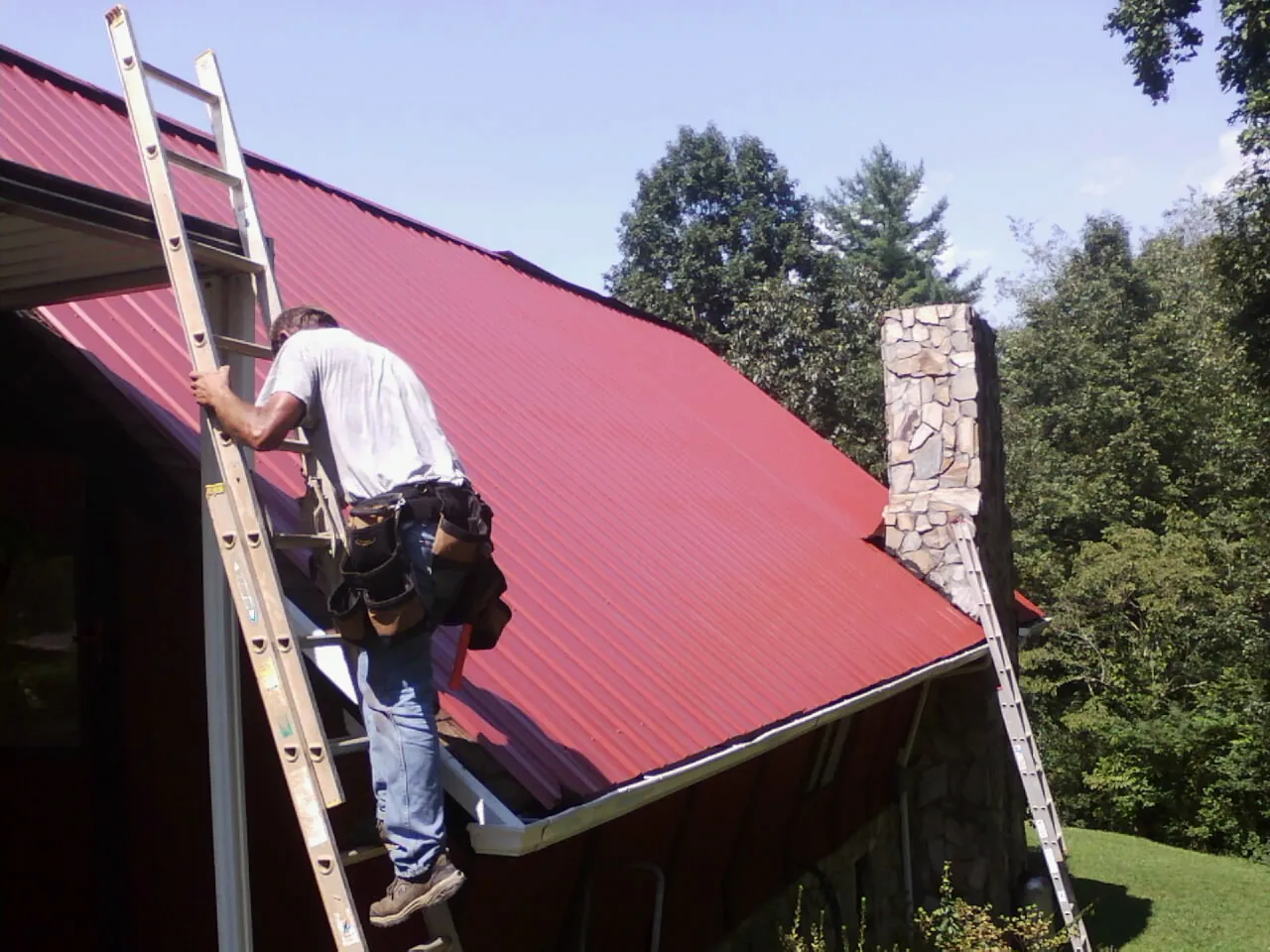 Roofer on a ladder beside a red metal roof and stone chimney — Soffit & Fascia Repair by Roofing Brothers in Pomeroy
